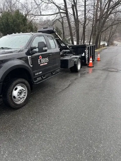 E'Bros pickup with a hook lift hauling a black dumpster on a road in Auburn MA