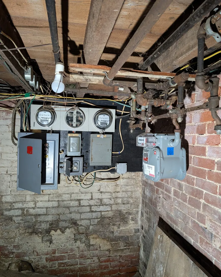Cluttered basement wall with old electric meters and disconnects mounted on brick before cleanup