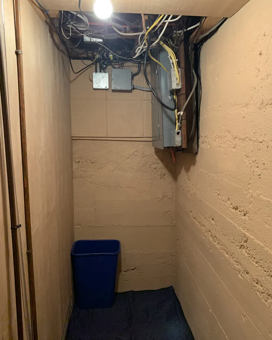 Cramped basement closet with an old electrical panel and overhead wiring before replacement