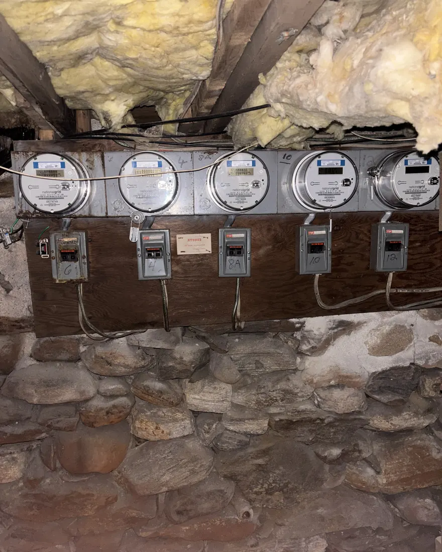 Five aging electric meters on a rotting wood backboard over a stone foundation wall before rebuild